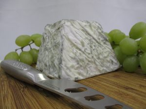 Valencay cheese is an example of a predominantly acid-coagulated cheese, sitting on a cheese board with a knife sitting with a knife and grapes in the background.