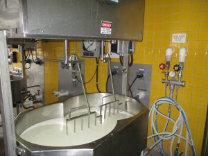 A photo of a double 'o' vat with automated cheese knives inserted in the vat of curd. The knives are attached to the top of the machine and rotate to cut the curd.