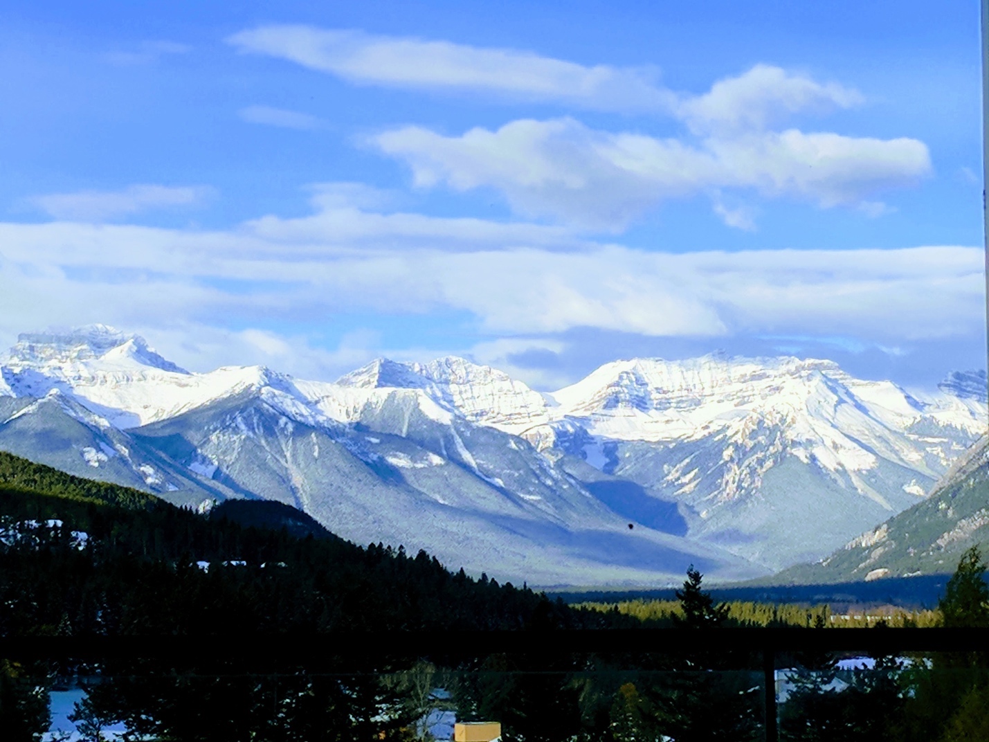 A long-shot of the Canadian Rocky Mountains. The sky is blue and a little cloudy; the mountains are snow-capped; the closer landscape includes lush green forest.]