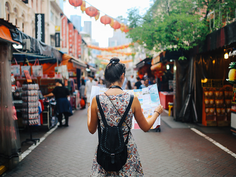 A young woman dressed casually with her dark hair in a bun walks through a Singapore street market wearing a backpack and reading a map.